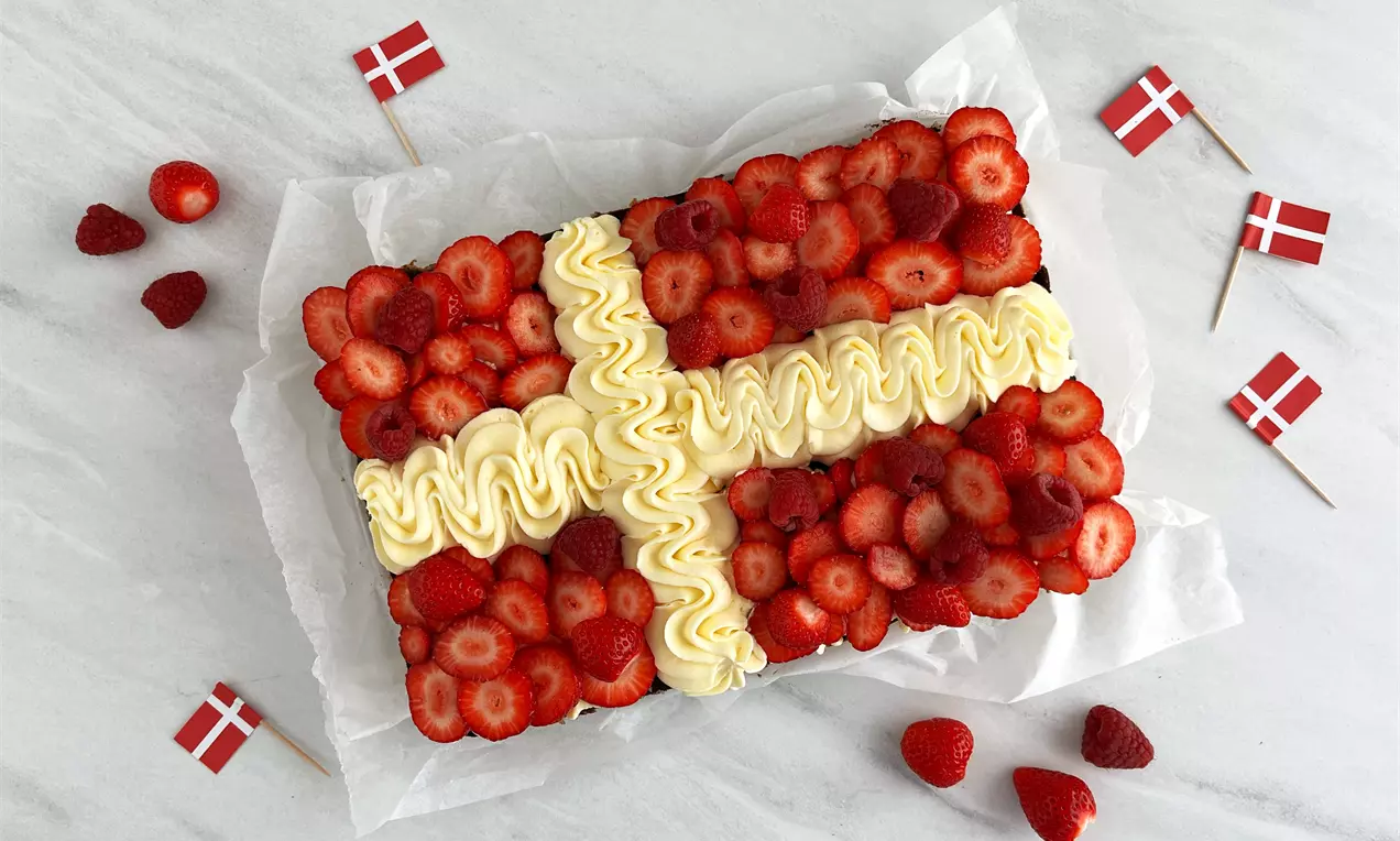 Rektangulær bradepandekage dækket med røde jordbær og flødeskumskryds som Dannebrogsflag, omgivet af små danske flag og bær på hvidt papir