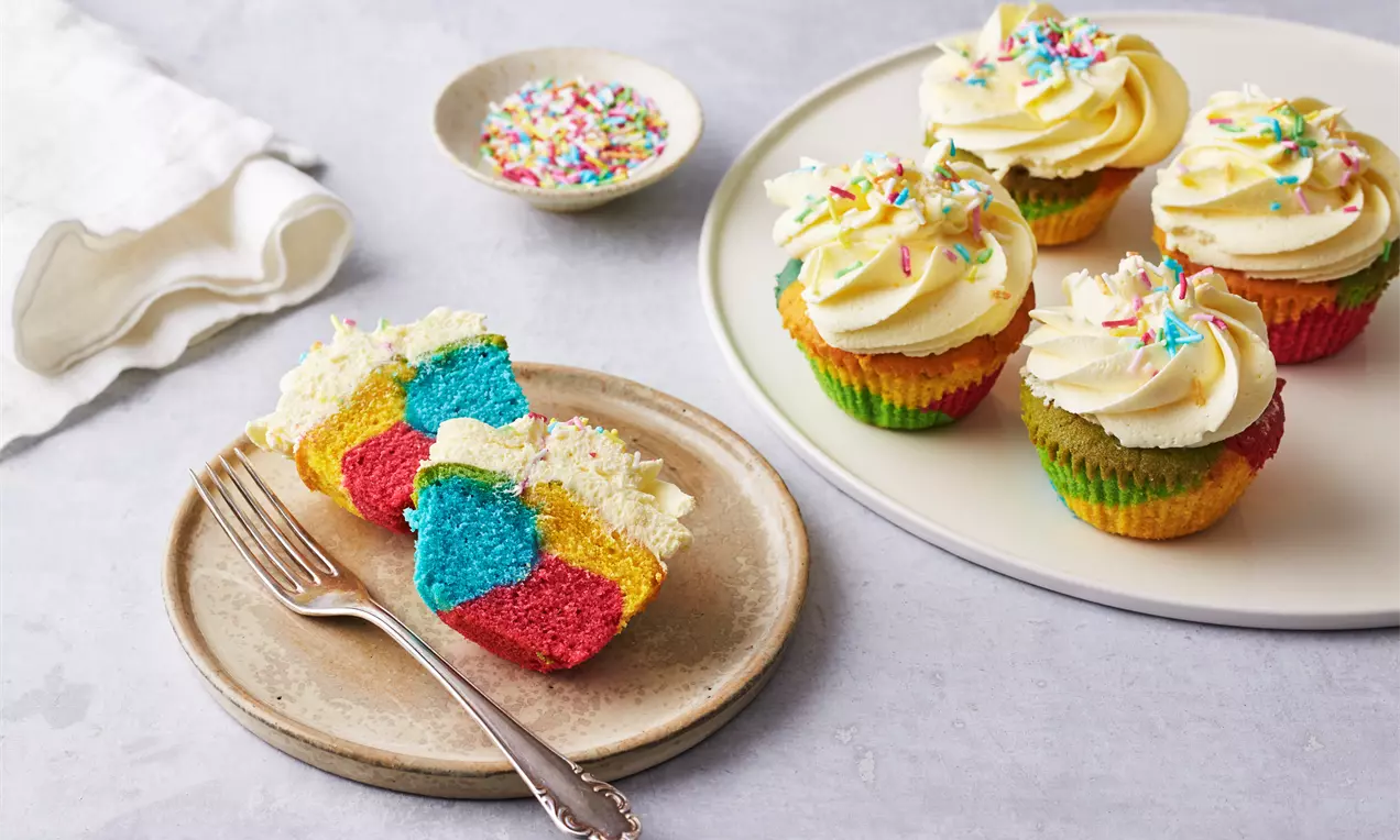 Farverige regnbuemuffins med lag i rød, blå og gul, toppet med hvirvlet hvid frosting og drys på tallerkener og i skåle