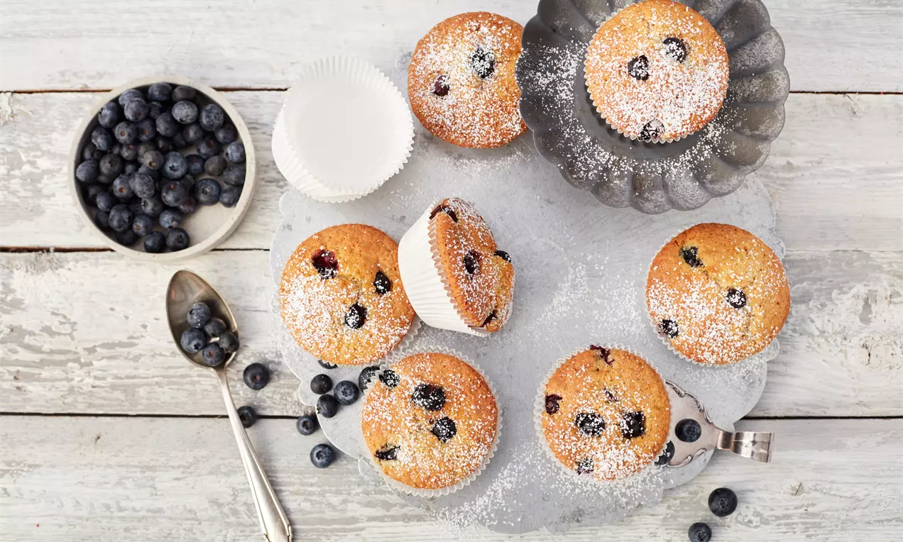 Glutenfri blåbærmuffins med gylden overflade drysset med flormelis, fordelt på et rustikt bord med løsrevne blåbær og en metalske