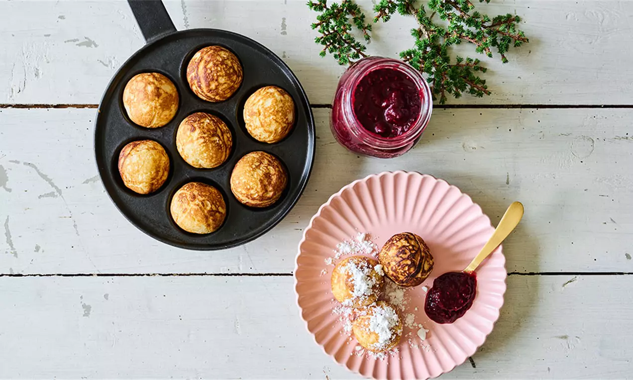 Gyldne æbleskiver med brunet overflade i sort pande ved siden af lyserød tallerken med æbleskiver, flormelis og rød marmelade