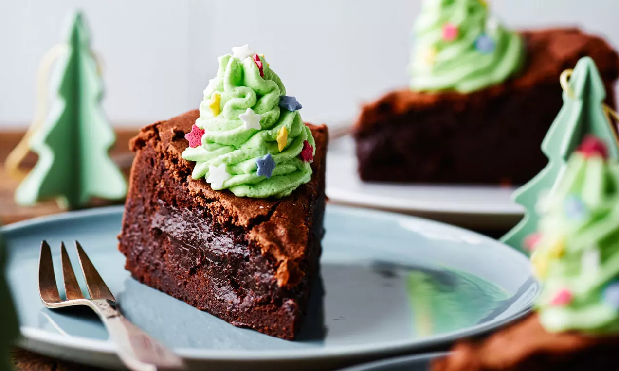 Chokoladekonfektkage med mørk brun, fugtig tekstur toppet med grøn frosting formet som juletræ og dekoreret med stjernefarvet krymmel på hvid tallerken