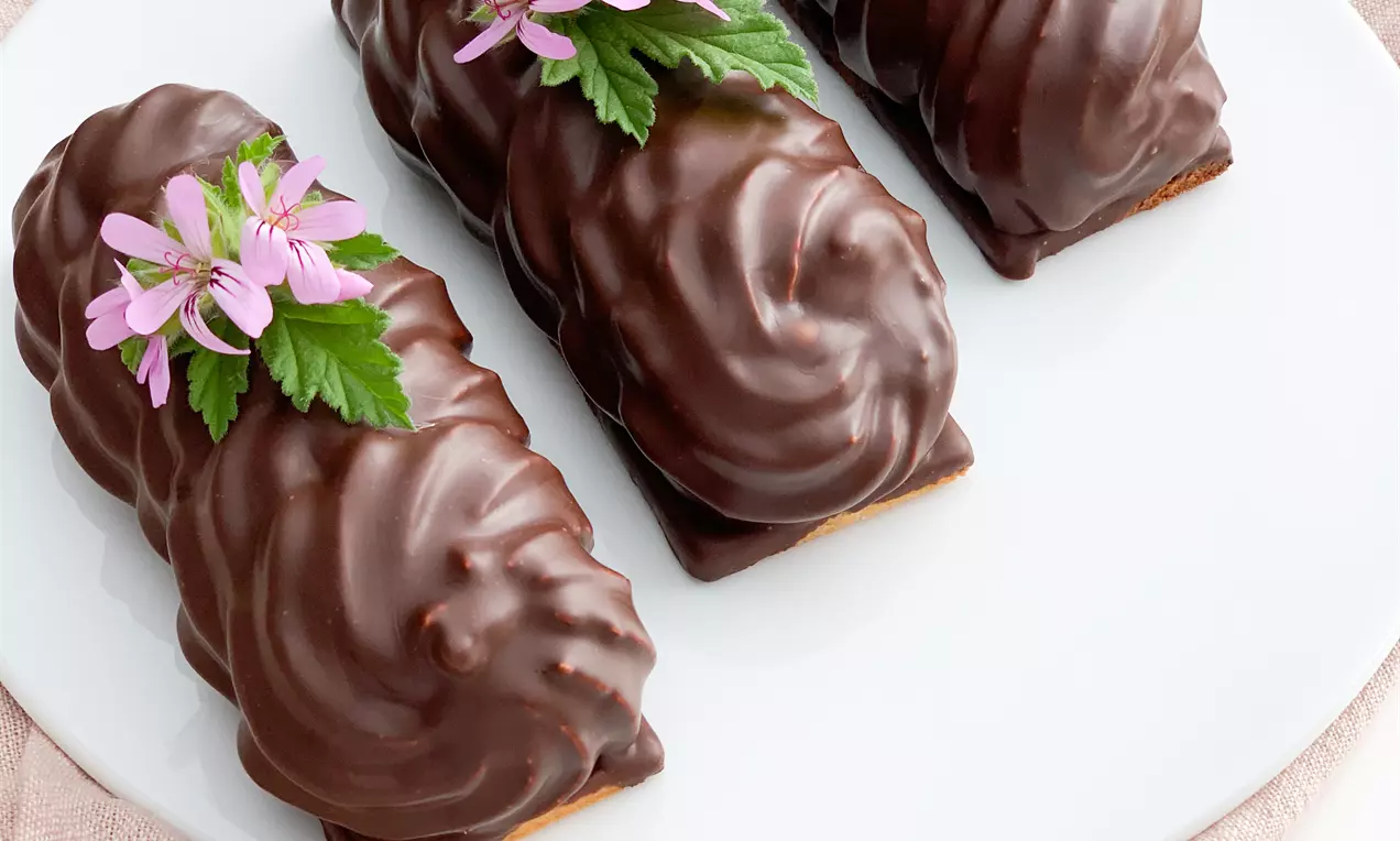 Tre aflange flødeboller med mørk, blank chokoladeoverflade pyntet med lyserøde blomster og grønne blade, serveret på en hvid tallerken