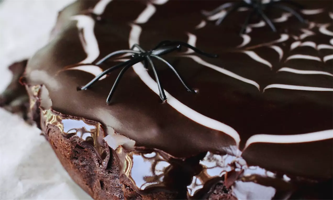 Chokoladekage med blank glasur dekoreret med hvidt spindelvævsmønster og sorte plastik-edderkopper på en lys baggrund