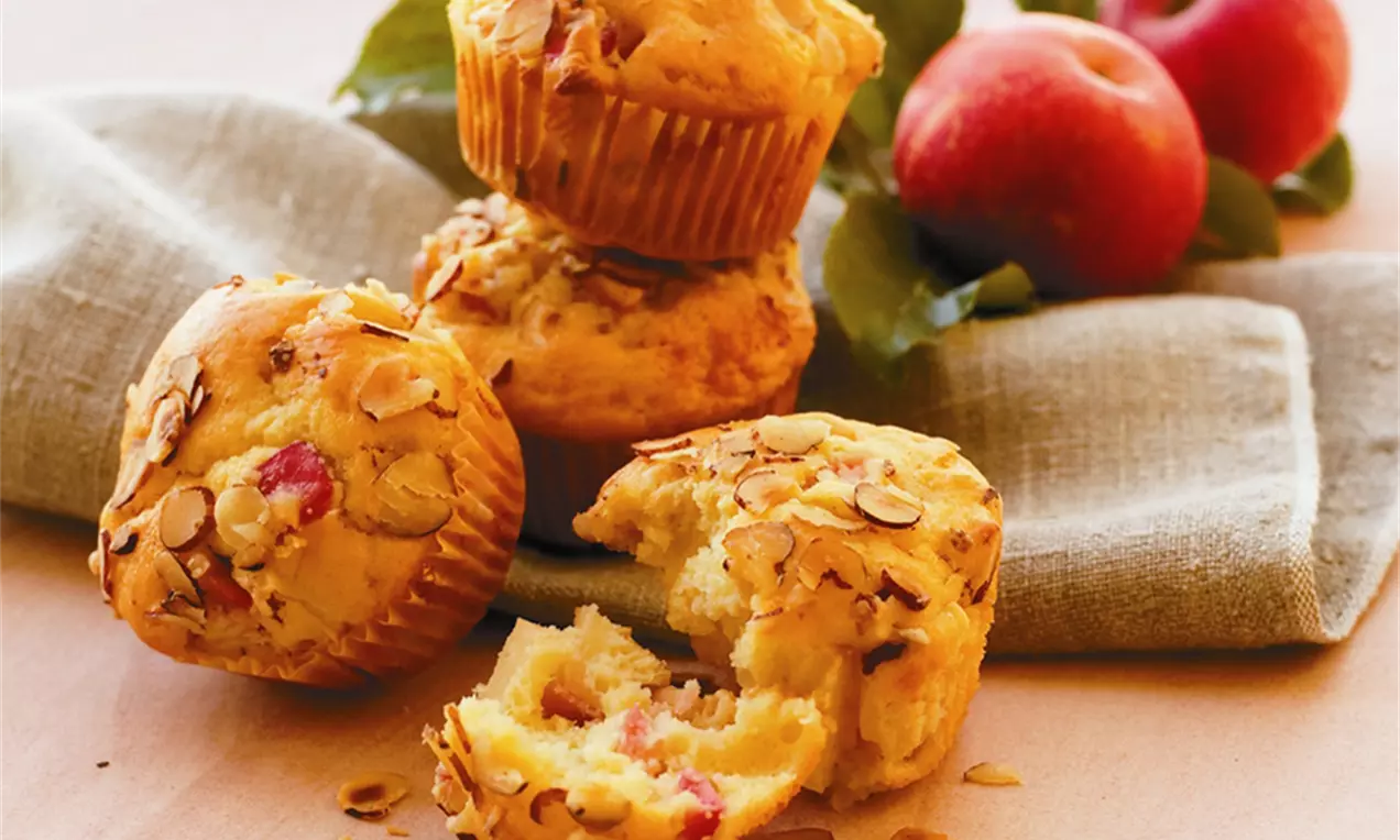 Gyldne æblemuffins med sprøde mandelflager, bløde stykker æble og let smuldrende tekstur på beige stof ved siden af røde æbler