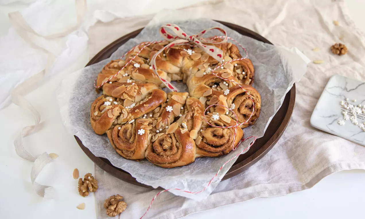 Kringle med gyldenbrune spiralformede kanelsnegle og valnøddestykker, drysset med hvide sukkerblomster på bagepapir og fad
