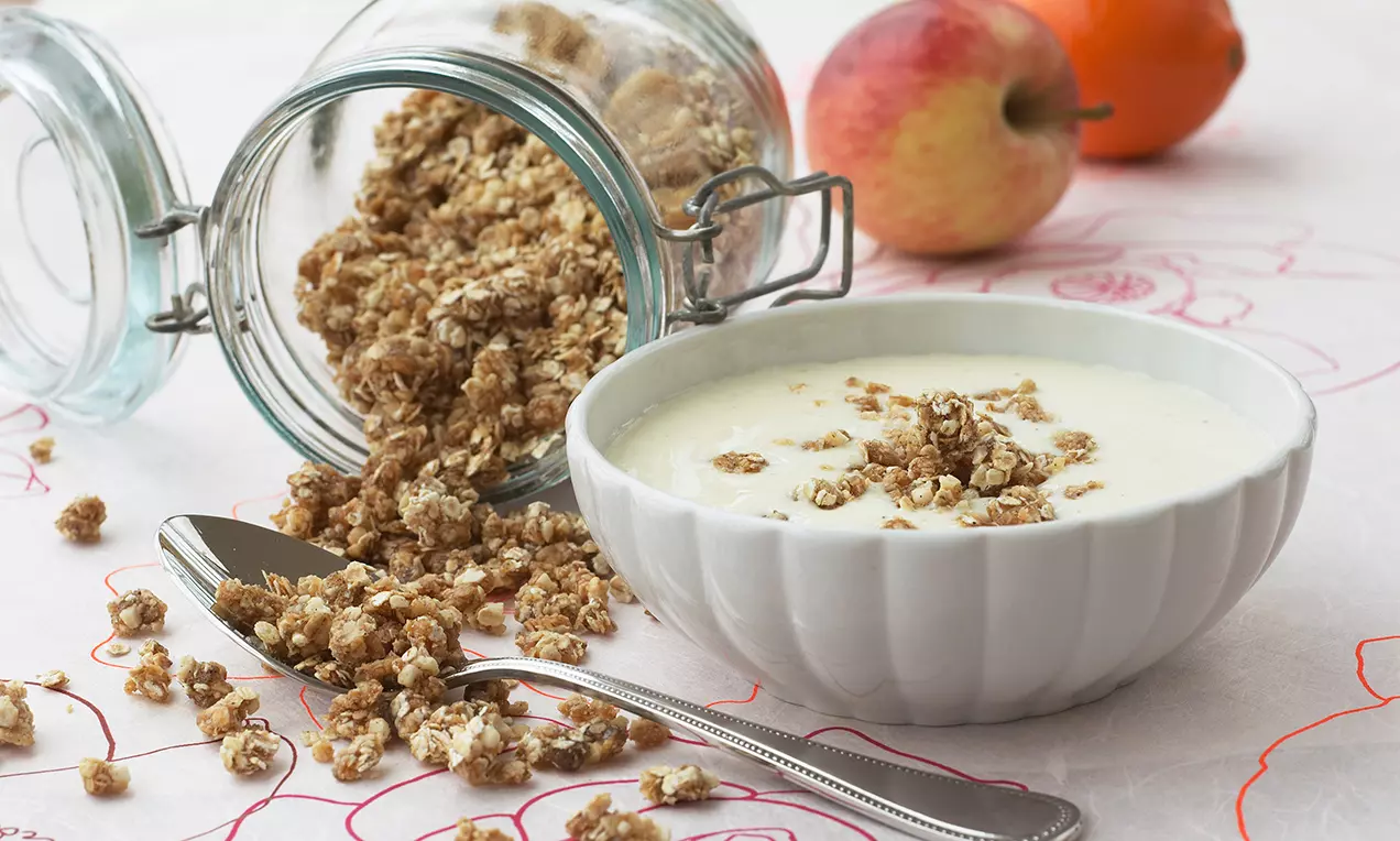 Hvid skål med lys koldskål toppet med gyldne sprøde knas på lys dug ved siden af et åbent glas med granola, ske og friske frugter