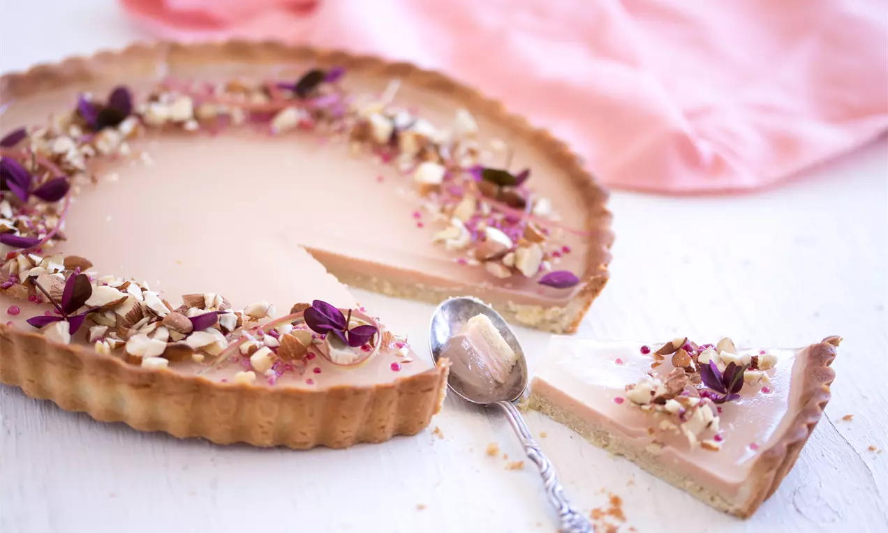 Lys rosa rabarbercreme på sprød, bølget tærtebund pyntet med hakkede nødder og små violette blomster på hvidt bord med sølvteske