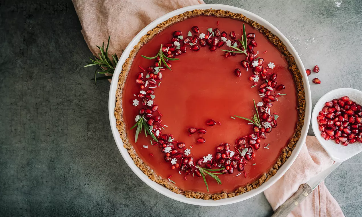 Julepanacottatærte med glat, rød overflade omkranset af brun kiksebund pyntet med gran, granatæblekerner og små hvide sukkerblomster