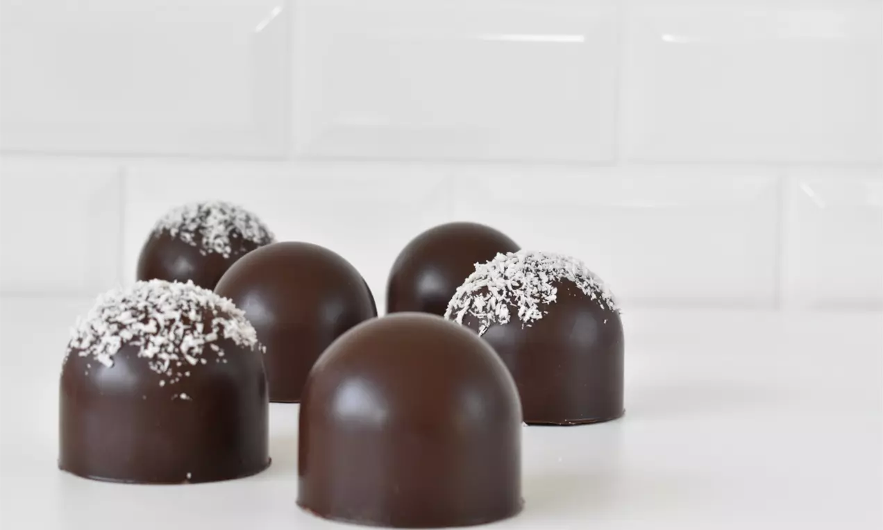 Flødeboller med glat mørk chokoladeoverflade, nogle drysset med hvid kokos, placeret på en hvid flade mod blanke hvide fliser