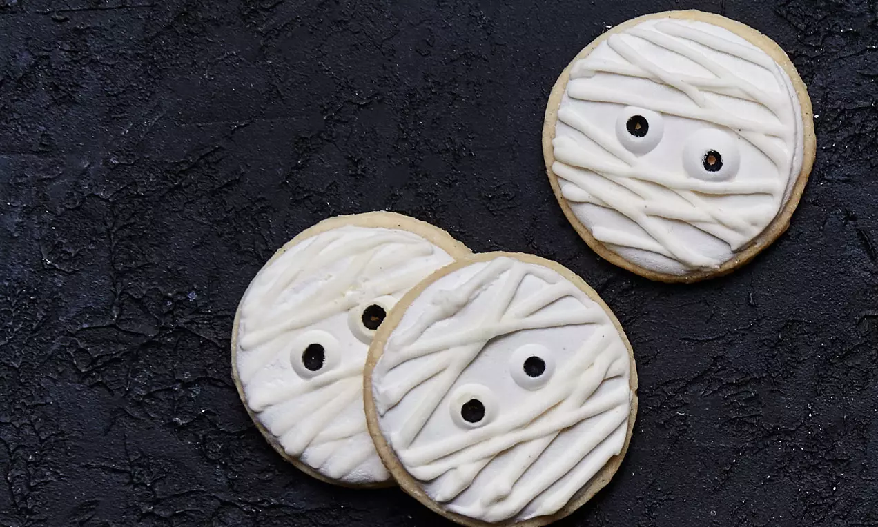 Runde cookies med lysebrun bund og hvid glasur formet som mumiebind, sorte sukkerojne placeret symmetrisk, liggende på sort ru baggrund