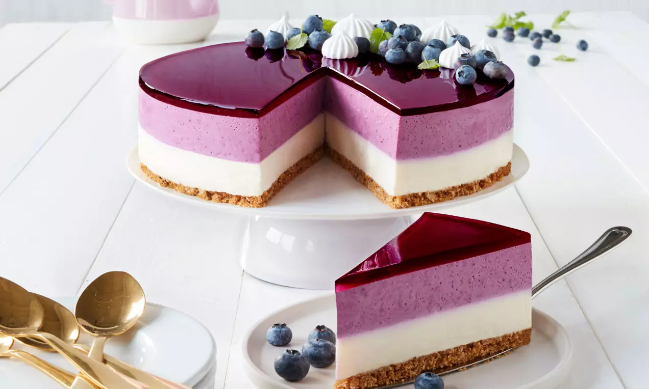 Cheesecake med gylden kiksebund, lag af hvid og lilla creme, mørklilla gelélag, pyntet med blåbær og flødeskumstoppe på hvidt fad