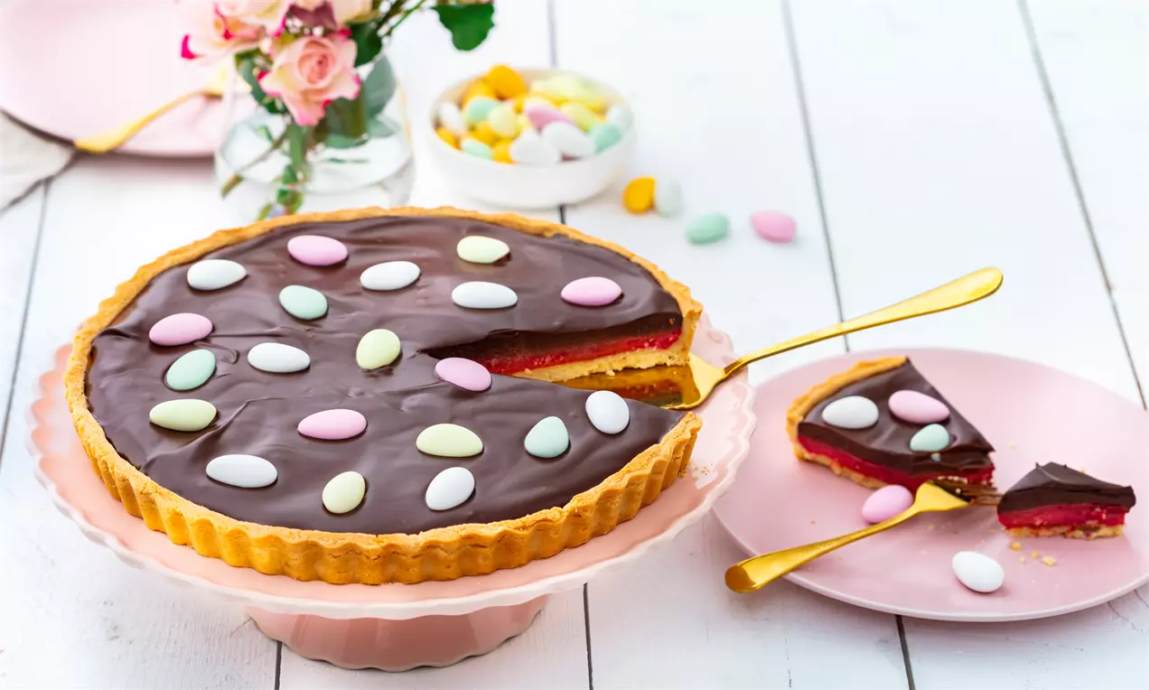 Påsketærte med blank mørk chokoladeganache og lyserød hindbærcurd, pyntet med farvede sukkeræg på et lyserødt kagefad