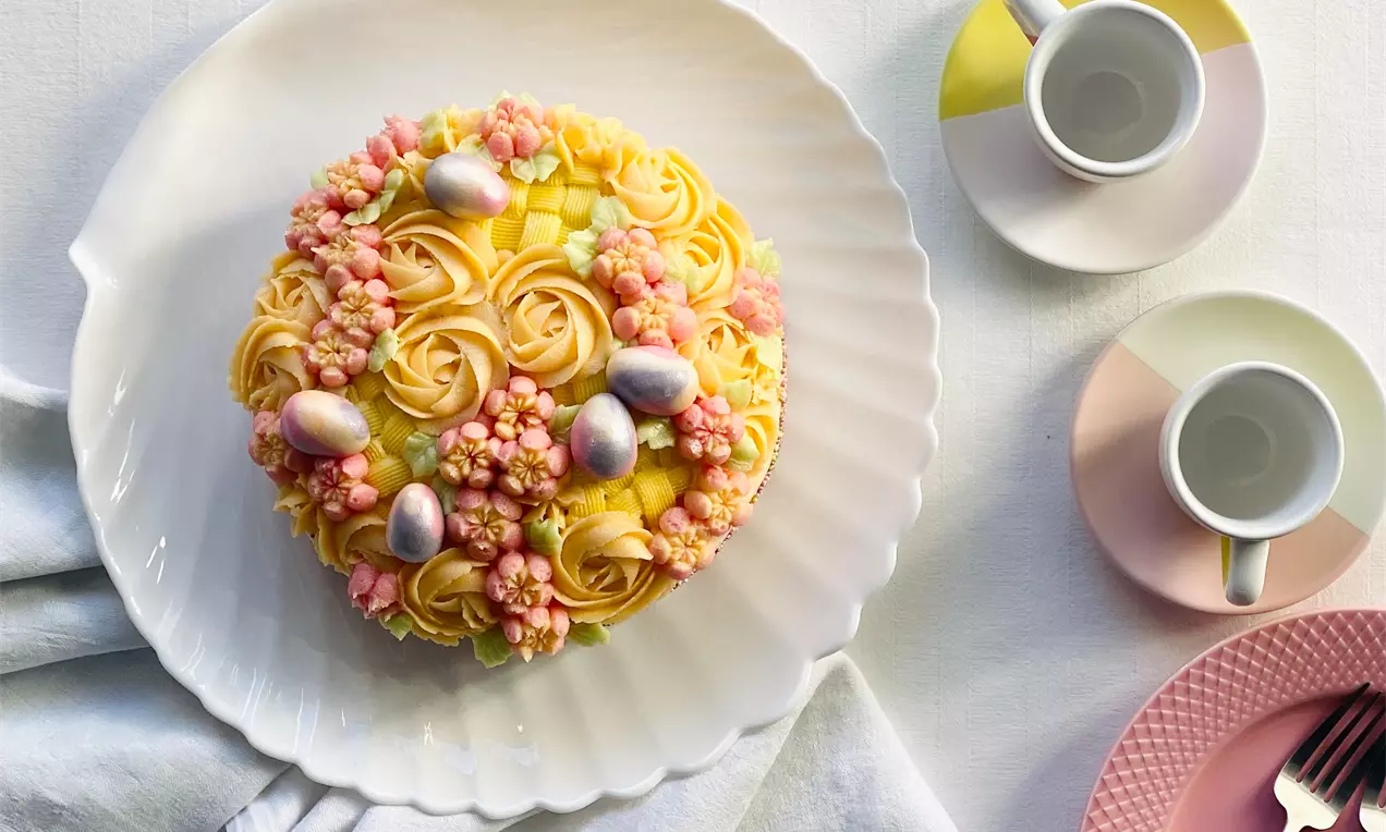 Påskekage med gule rosetter og små lyserøde blomster, sølvæg og ternede grønne detaljer på en hvid rillet tallerken