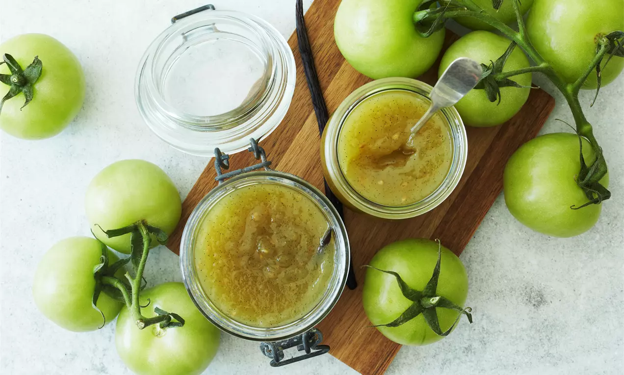 Grøn tomatmarmelade med blank, syltet tekstur i åbent sylteglas på træbræt omgivet af friske lysegrønne tomater med stilke