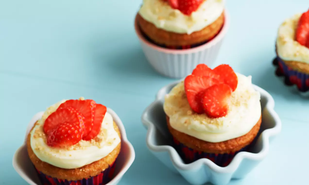 Jordbær-cheesecake-cupcakes med gyldenbrun bund, cremet hvid topping og friske røde jordbær, serveret i små formede skåle på lyseblå baggrund