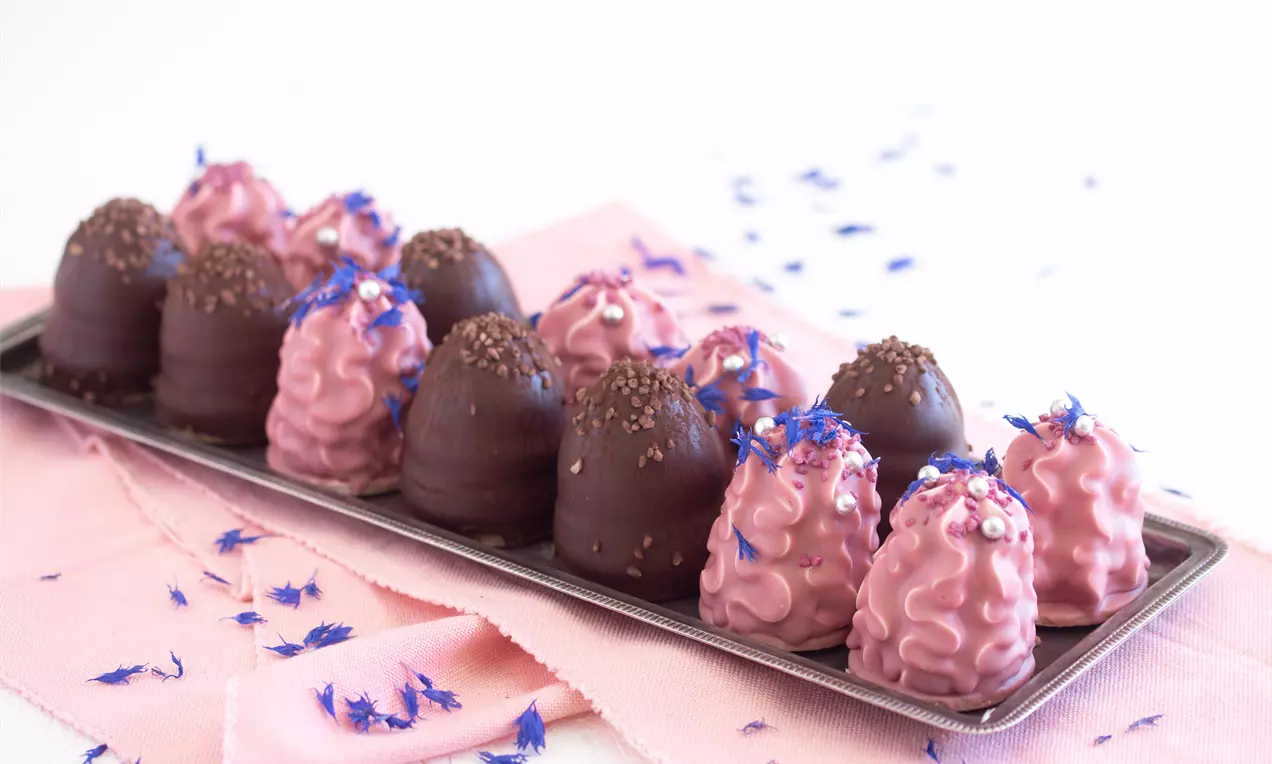 Flødeboller med pink og brun glasur, enkelte pyntet med blå blomster på en metalbakke, liggende på lyserosa stof med spredte blomster