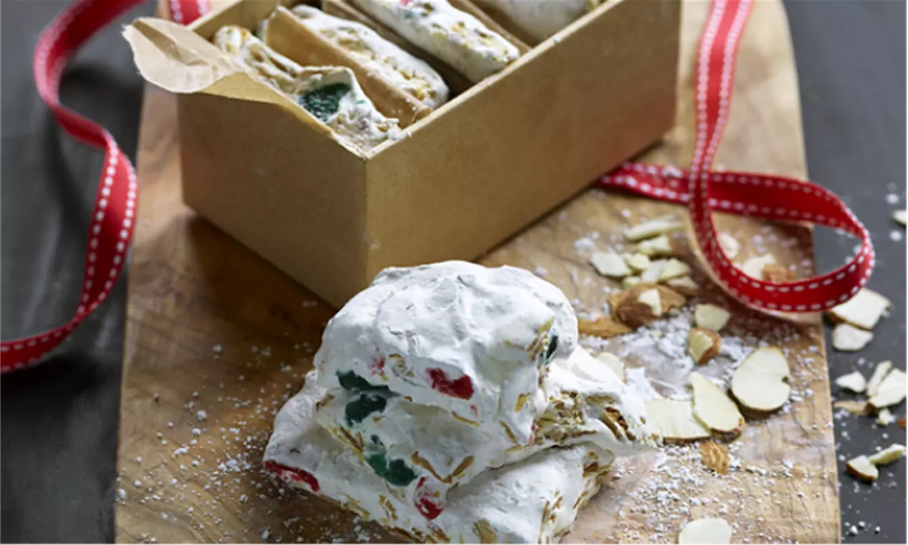 Fransk nougat med skinnende, røde og grønne cocktailbær og gyldne ristede mandler, indpakket i hvidt konfekt lagt på træbræt