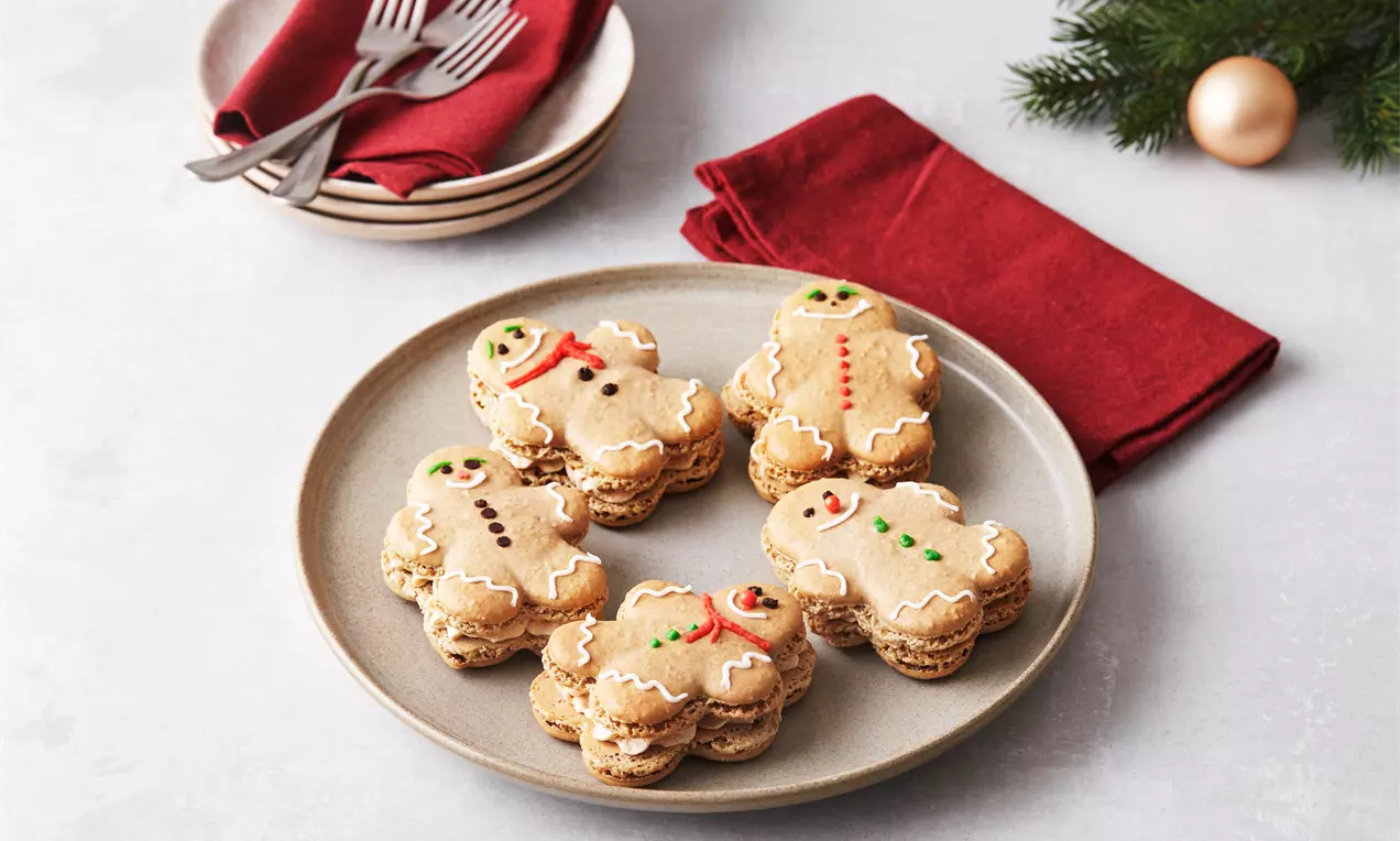 Honningkageformede macarons med lys brun farve og hvid glasur, fyldte med creme, placeret på en beige tallerken med rødt stof og julepynt