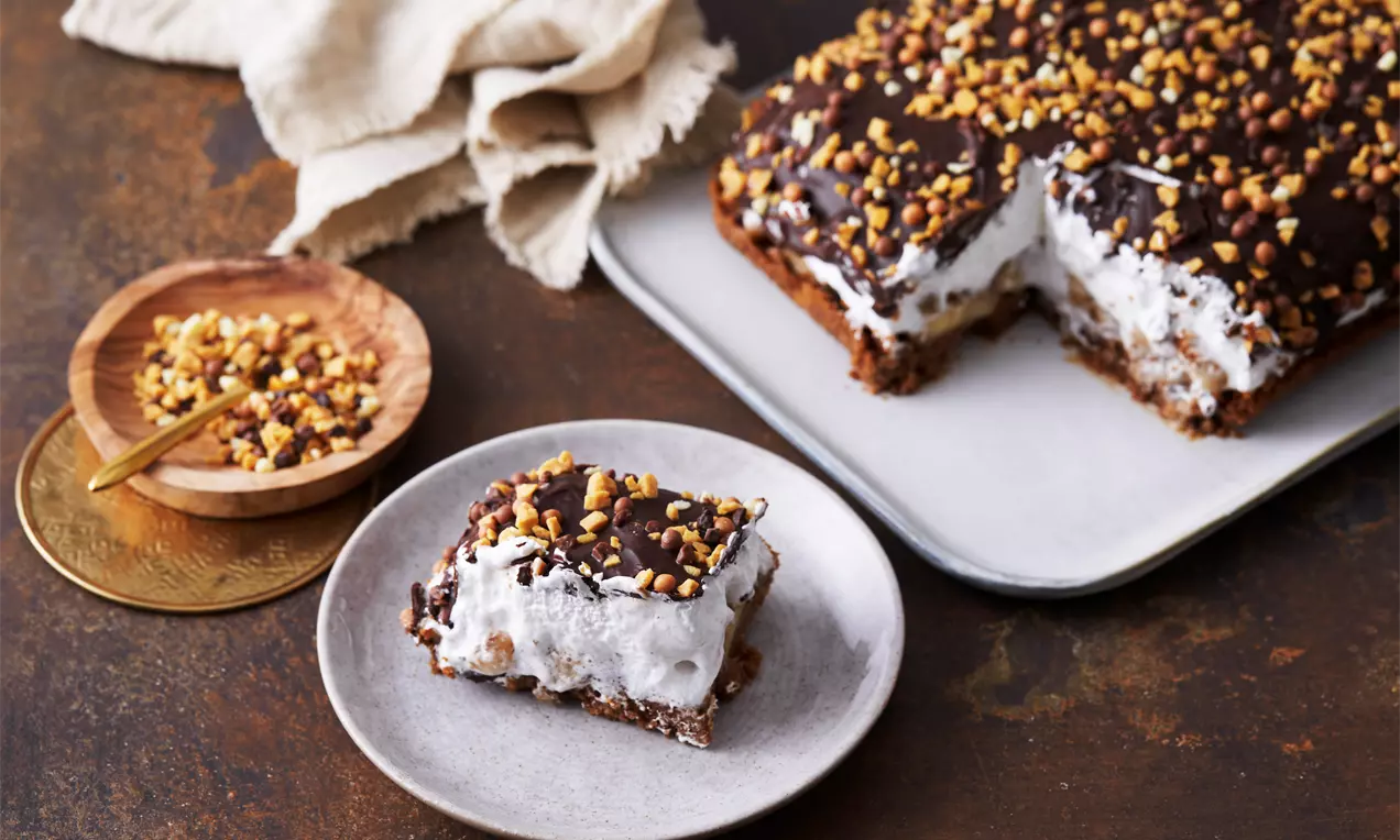 Banoffee flødebolle-kage med luftig hvid skum fyld på brun kiksebund, toppet med mørk chokolade og gyldne hasselnødstykker på en lys tallerken
