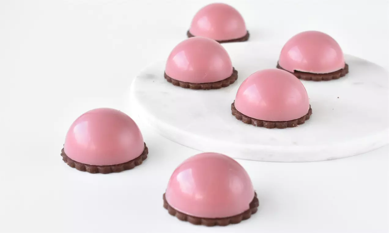 Ruby-flødeboller med glat lyserød overflade på sprøde chokoladebunde placeret dels på hvidt marmorfad, dels på hvid baggrund