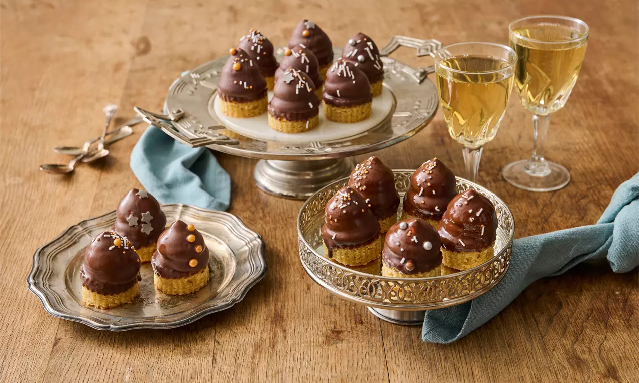 Små flødebolle petit fours med mørk chokoladeovertræk og pynt på gylden kiksebund anrettet på sølvfade ved et træbord med glas og servietter