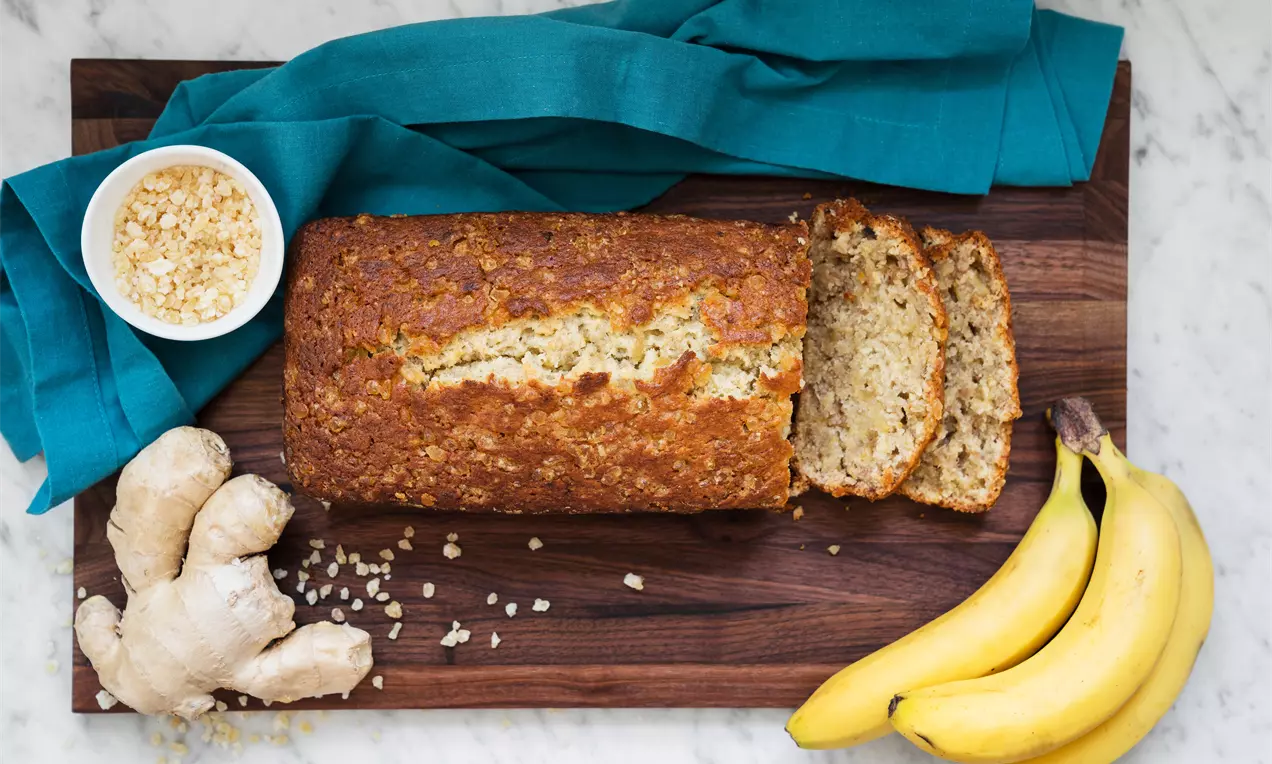 Gyldenbrun bananbrød med saftig tekstur og synlige ingefærstykker på mørkt træbræt, omkranset af friske bananer, ingefær og blå stofserviet