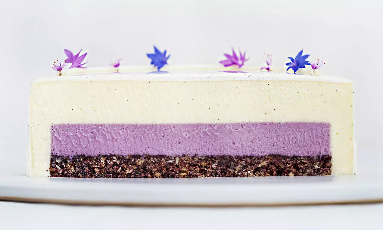 Rektangulær kage med lysegul vaniljeglaze, lyse-lilla blåbærmousse og mørk bund, pyntet med små violette blomster på toppen
