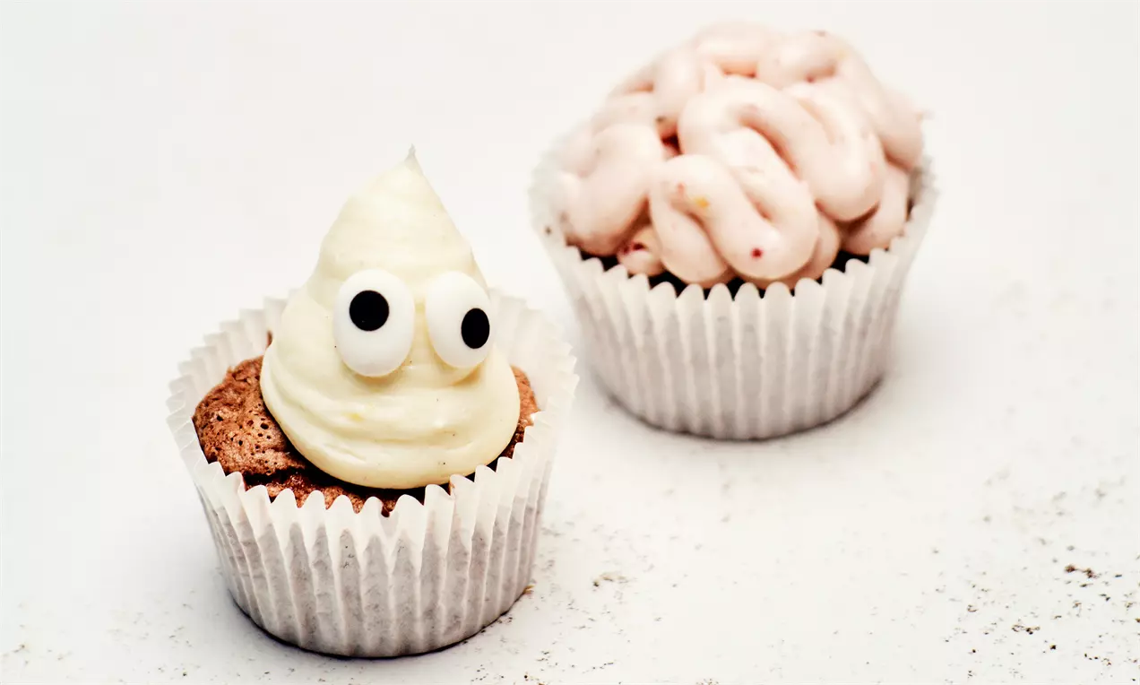 Græskar-muffins i hvide papforme, den ene toppet med hvid frosting og sorte øjne, den anden med lyserød hjerneformet pynt
