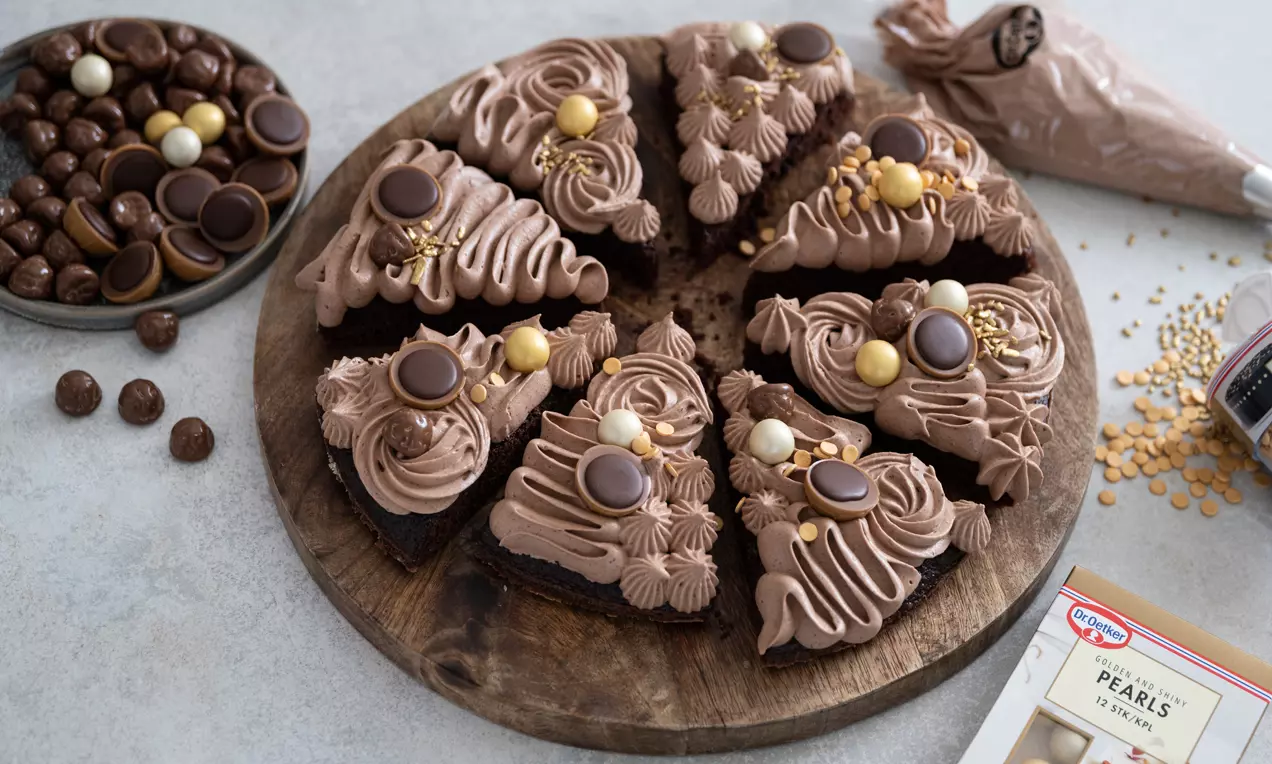 Chokoladekage skåret i trekanter, mørkebrune med blød nougatfarvet frosting og pyntet med hasselnøddekorationer på træfad