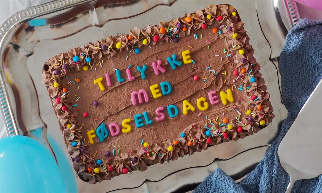 Firkantet bradepandekage med brun chokoladecreme og farvede bogstaver, kantpyntet med chokolade og krymmel på sølvfad