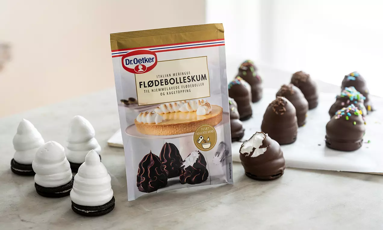 Hvide flødeboller med glat skum på mørke kakaobunde samt chokoladeovertrukne flødeboller med drys på en lys bordplade ved siden af en produktpakke