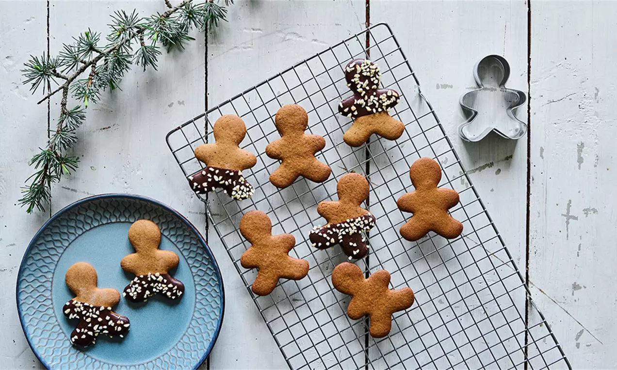 Brunkagemænd gyldenbrune med chokoladedyppede arme og hvid krymmel ligger på en sort bagerist og blå tallerken ved siden af en grangren