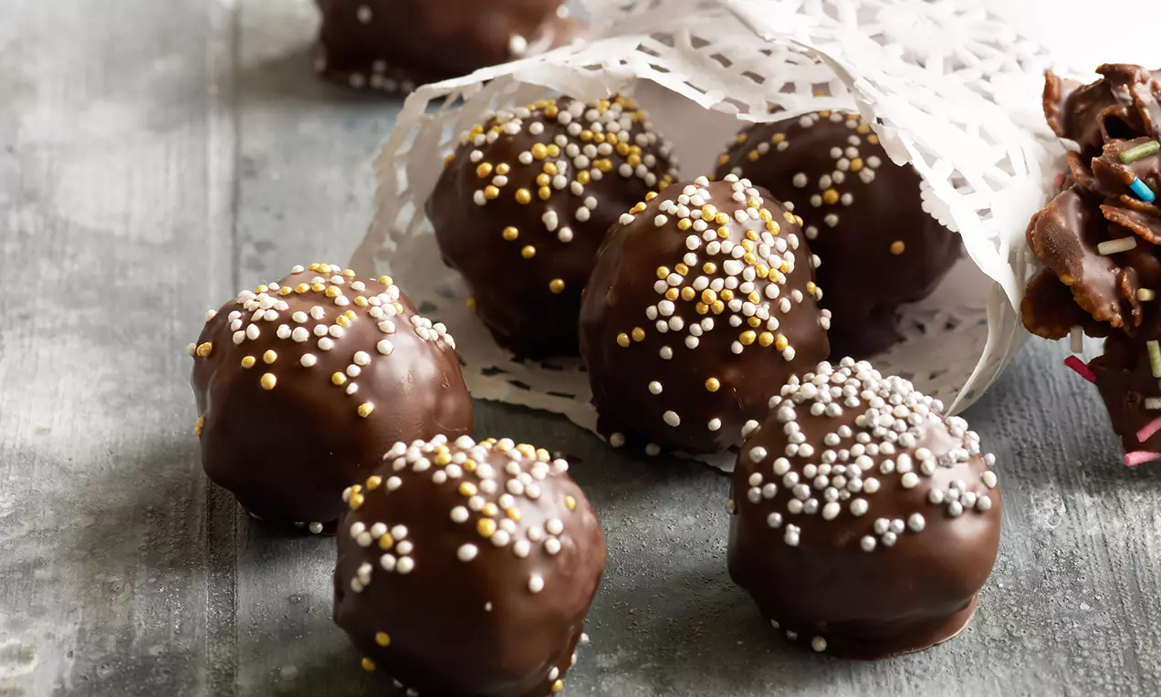 Små kugler dækket af mørk chokolade og pyntet med farvede sukkerperler, liggende på en grå overflade ved siden af hvid papirpynt