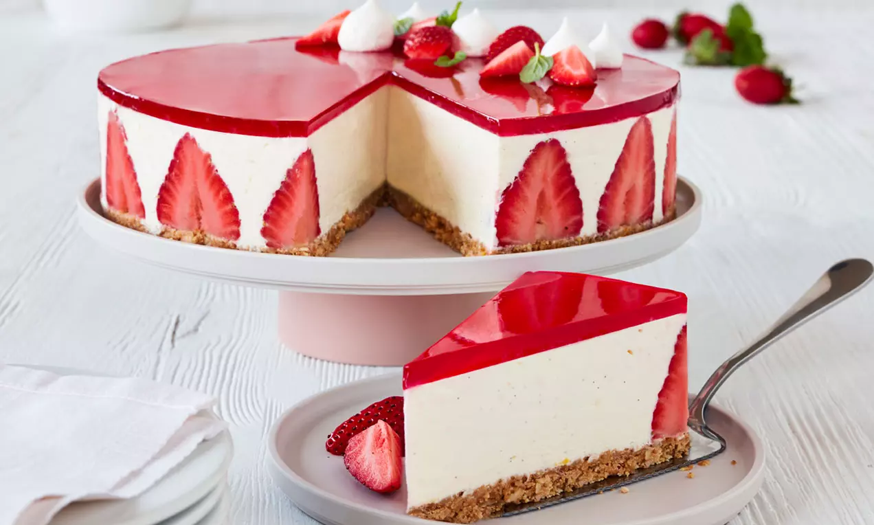 Cheesecake med lysebrunt kiksebund og cremefarvet fyld dekoreret med røde jordbærskiver og spejlblank jordbærgelé på hvidt kagefad