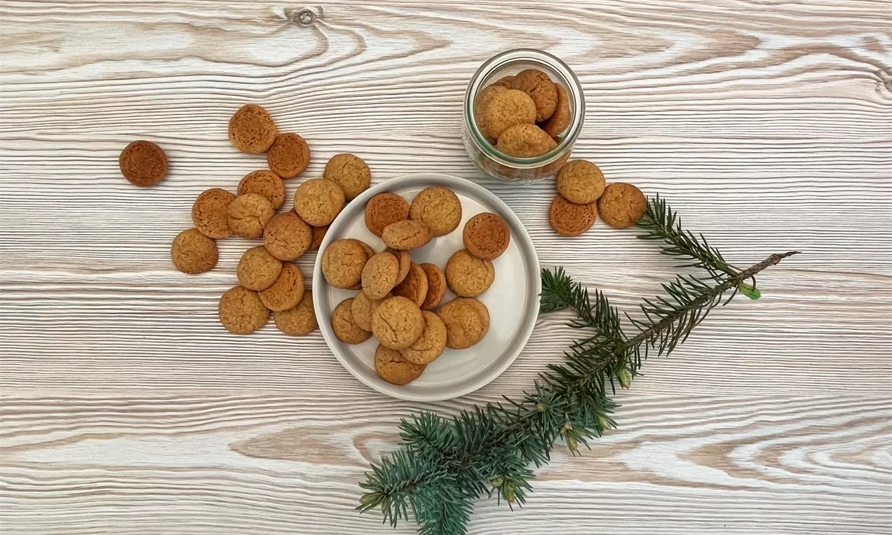 Små, runde, gyldne veganske pebernødder med let grov tekstur spredt på og omkring en beige tallerken med grangren og glas på lys træbord