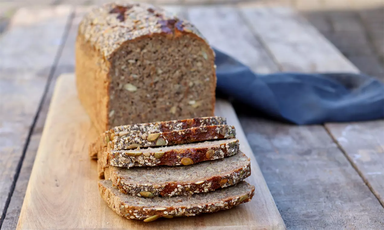 Rugbrød med kerner og sprød skorpe skåret i skiver på et lyst træbræt, omgivet af et mørkt stof på et rustikt bord