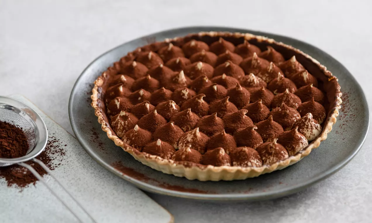 Tiramisu-tærte med gyldne mascarponetoppe drysset med brun kakao, serveret på en grå tallerken ved siden af en sigte med kakaopulver