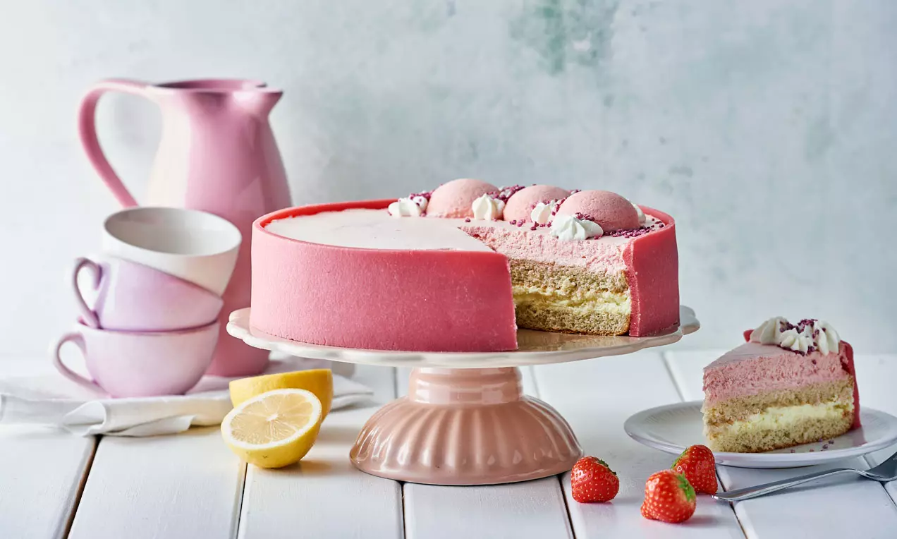 Lyserød sommerlagkage med glat overflade og lyse bundlag på lys rosa fad omgivet af friske jordbær, porcelæn og en citron