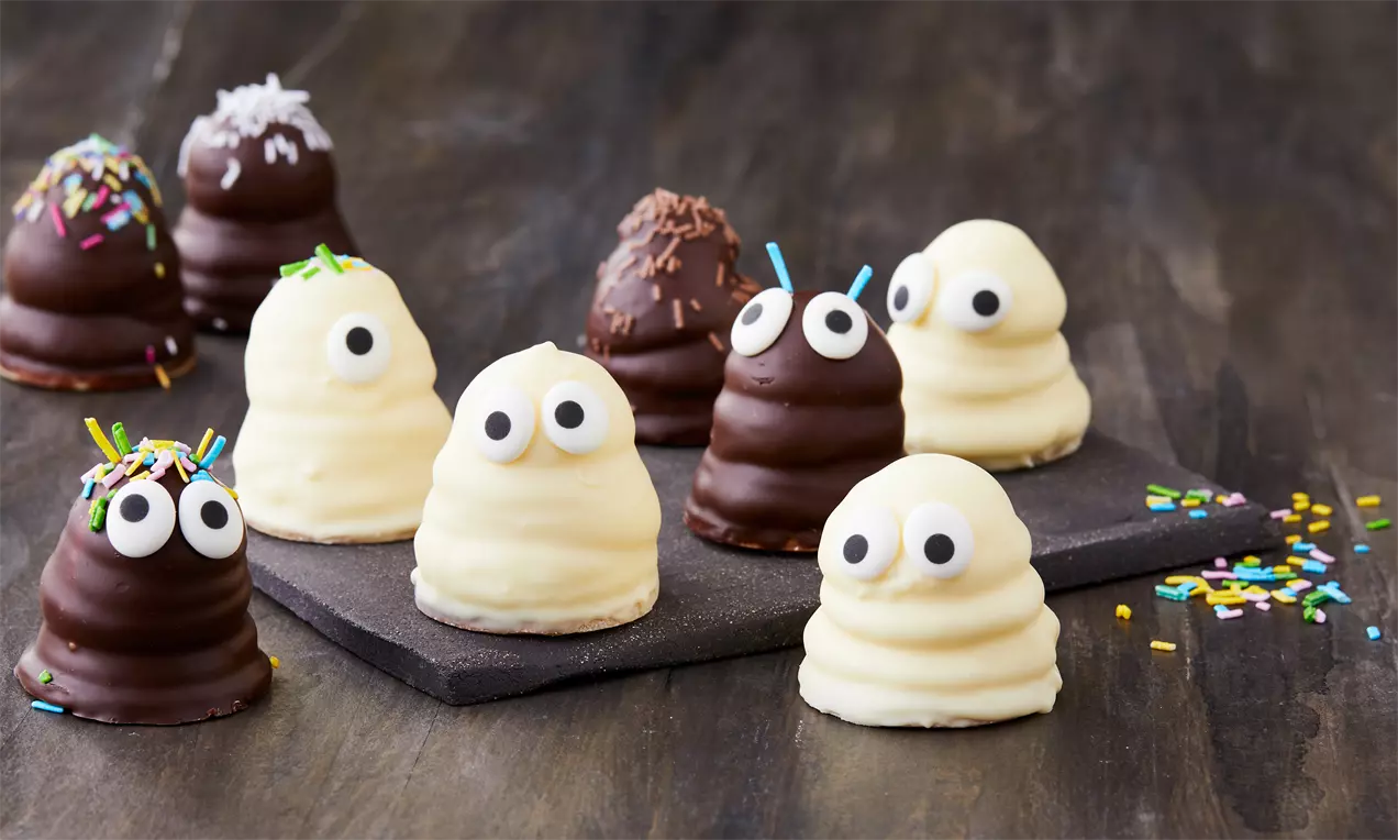 Små flødeboller formet som spøgelser og monstre med hvide og chokoladefarvede overtræk, pyntet med øjne og krymmel på en mørk træoverflade