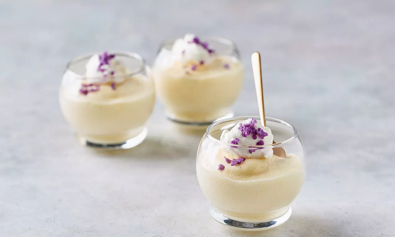 Lys gul citronfromage med blød tekstur serveret i runde, klare glas toppet med hvid creme og små lilla sukkerblomster