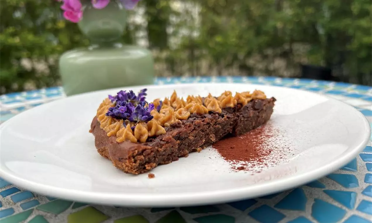 Vegansk kikærte-brownie med mørk brun overflade og gyldne dadelkarameltoppe, pyntet med lilla blomster på en hvid tallerken